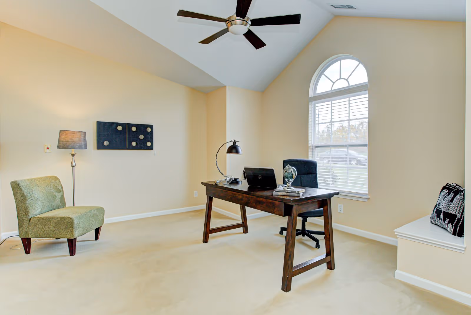 A bright office room with beige walls and carpet, featuring a wooden desk with a laptop, desk lamp, globe, and books. There is a black office chair behind the desk, a green upholstered chair to the left, a floor lamp, wall art, and a large arched window with white blinds letting in natural light. A bench with a decorative pillow is positioned under the window.