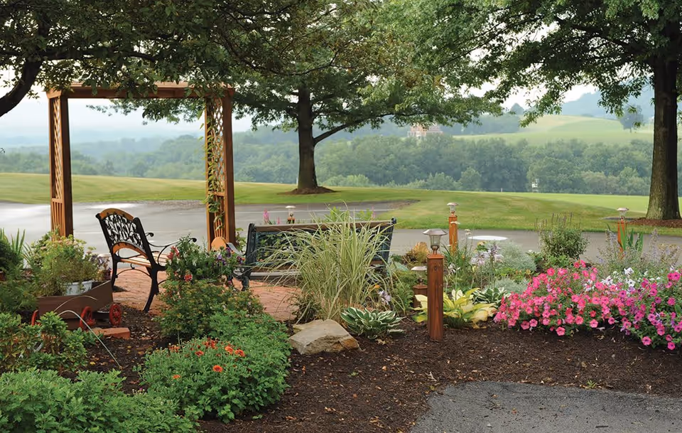 A peaceful outdoor garden area with a wooden pergola, two benches, and various flowering plants and shrubs. In the background, there are large trees and a scenic view of rolling green hills under a cloudy sky.