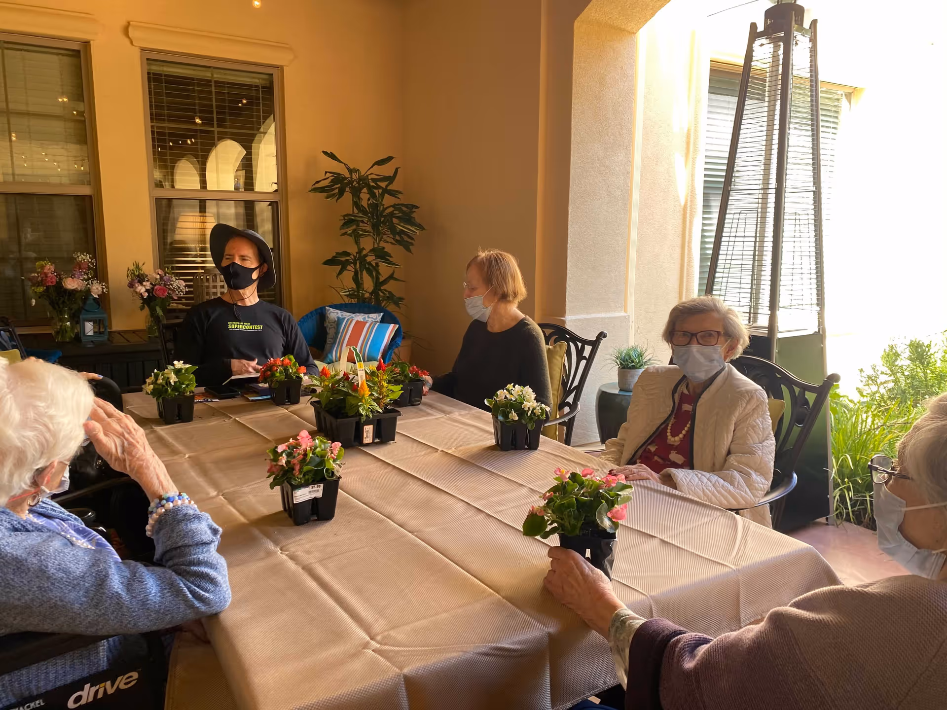 A group of elderly people wearing face masks sitting around a rectangular table covered with a beige tablecloth. Small potted flowering plants are placed on the table. The setting appears to be a covered outdoor patio area with windows, plants, and a tall outdoor heater visible in the background.
