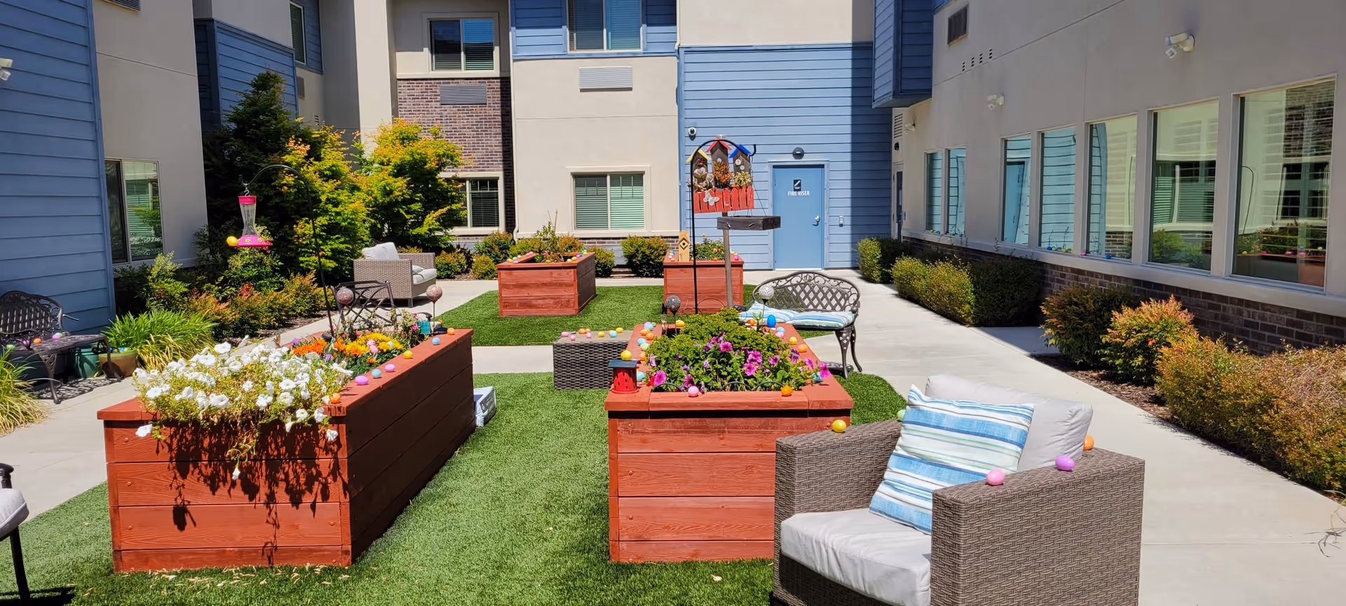 Outdoor courtyard area at Summerset Senior Living, Lincoln - Memory Care & Adaptive Care, featuring raised wooden garden beds with colorful flowers, green artificial turf, several cushioned chairs and benches, and a bird feeder hanging near the plants. The courtyard is surrounded by the building walls with windows and a blue door labeled 'Fire Hose'.