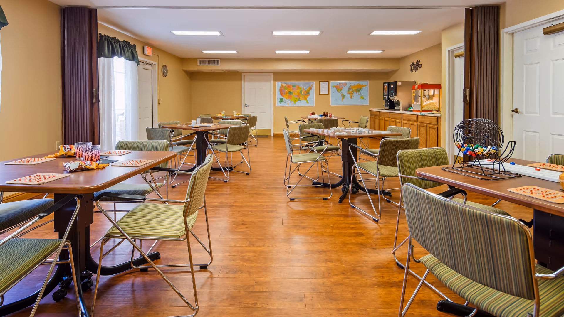 A well-lit activity room with multiple tables and green striped chairs arranged around them. The tables have bingo cards, markers, and snacks on them. There is a bingo cage with colorful balls on one table. The room has wooden flooring, beige walls, two maps on the far wall, and a counter with a popcorn machine and coffee dispensers. A door and windows with dark curtains are visible.