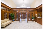 Spacious wood-paneled lobby with tiled floor, bench seating, decorative plants, and glass-paneled double doors beneath a vaulted ceiling.