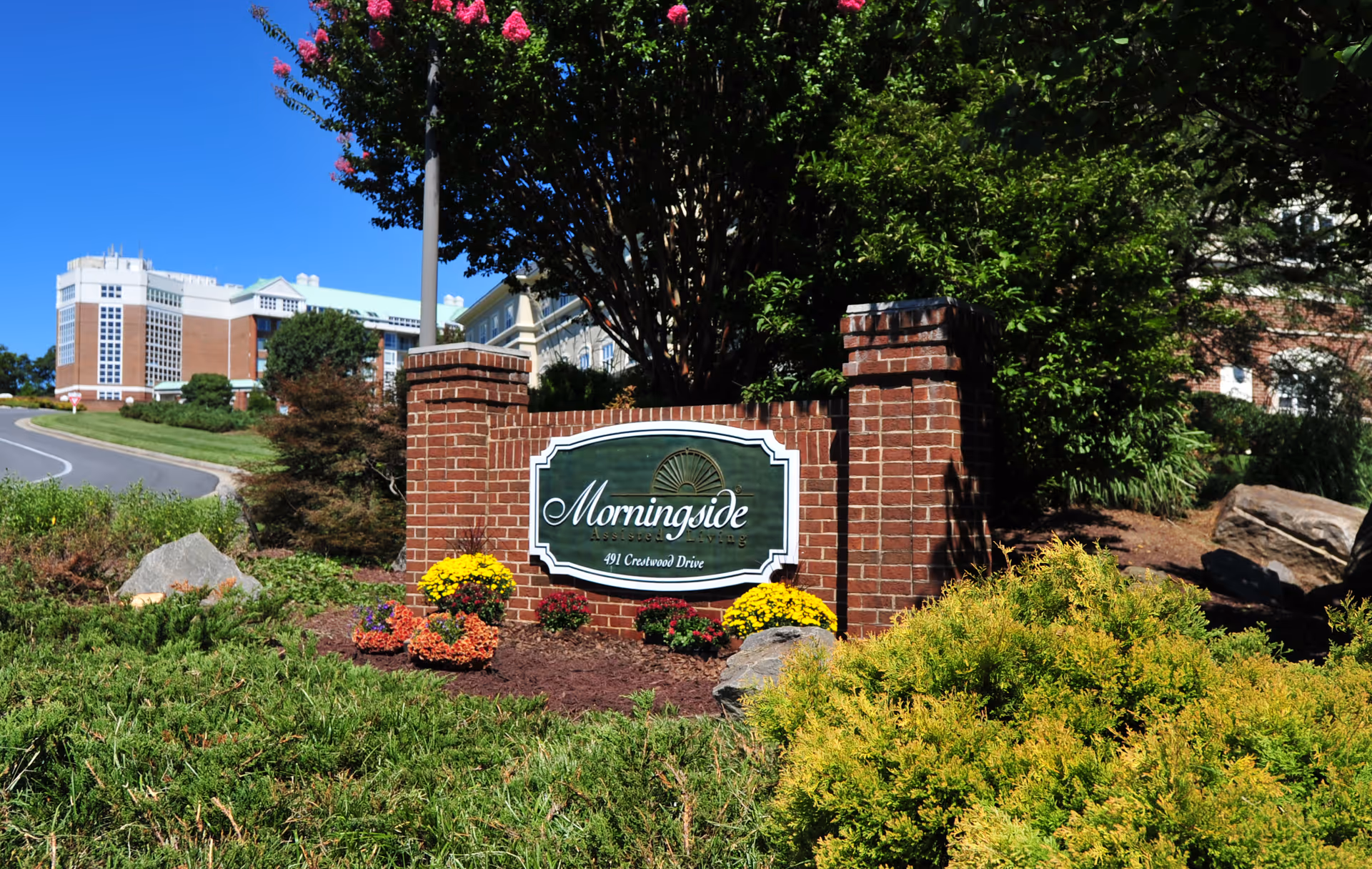 Brick sign with the name 'Morningside' and address '491 Crestwood Drive' surrounded by colorful flowers and greenery, with a large building and a clear blue sky in the background.