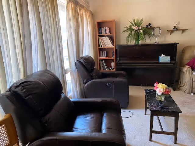 A cozy living room with two black leather recliners facing a small black marble coffee table with a flower arrangement. Behind the recliners is a window with sheer curtains allowing natural light to enter. In the background, there is a black upright piano with a flower vase on top, a wooden bookshelf filled with books, and a beige armchair partially visible on the right.