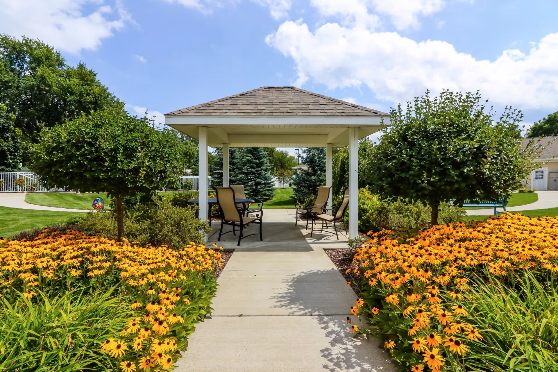 A garden pathway leading to a covered gazebo with chairs arranged underneath. The pathway is flanked by vibrant yellow and orange flowers and small manicured trees. The sky is partly cloudy with blue patches visible.