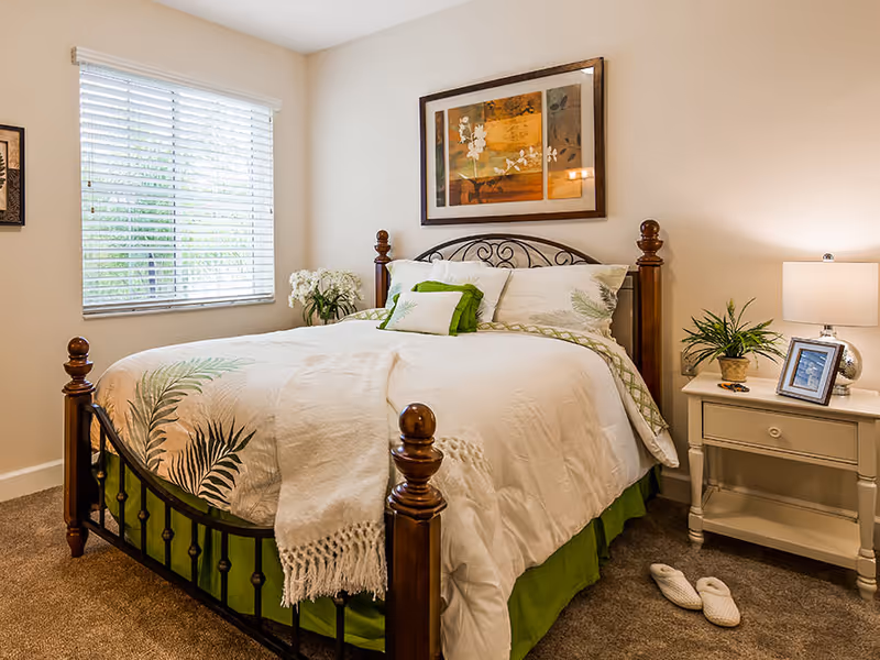 Cozy bedroom featuring a wooden bed with white and green bedding, a nightstand with a lamp and plant, and a window with blinds.