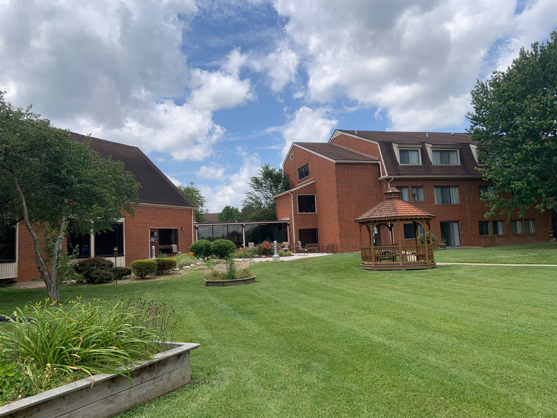 Outdoor view of a senior living facility with red brick buildings, a well-maintained green lawn, a wooden gazebo, trees, and a partly cloudy sky.