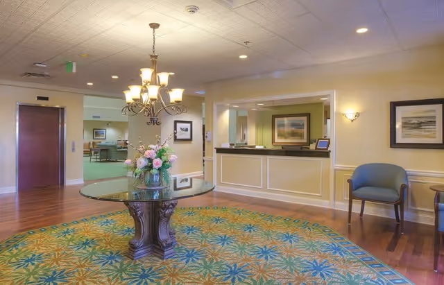 Interior view of a senior living facility lobby area with a round glass table holding a floral arrangement in the center on a patterned carpet. There is a chandelier above the table, a reception desk to the right, a blue chair, framed artwork on the walls, and an elevator door in the background.