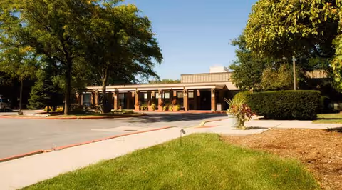 Exterior view of a single-story senior living facility building with a covered entrance, surrounded by trees, shrubs, and a well-maintained lawn under a clear blue sky.