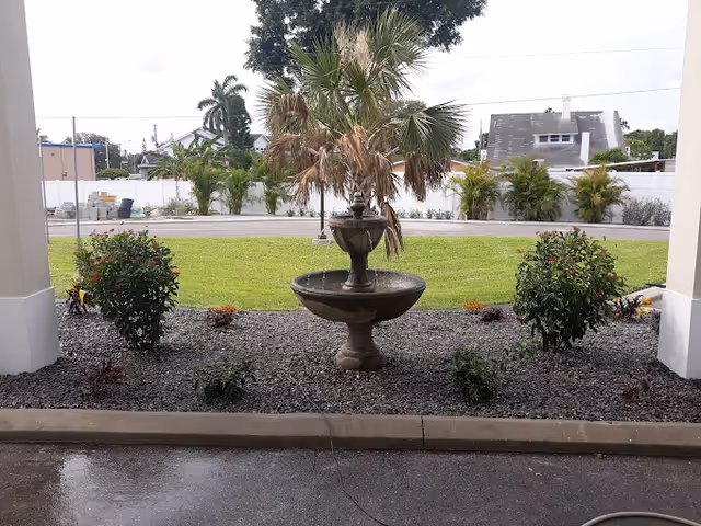 View of an outdoor garden area with a two-tiered stone fountain in the center, surrounded by small bushes and plants. Behind the garden is a green lawn, a white fence, and some houses and trees in the background.