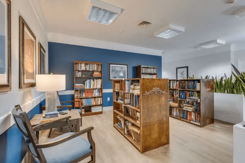 Interior view of a cozy reading room with wooden bookshelves filled with books, two wooden chairs with blue cushions, a small side table with a lamp and books, blue and white walls, framed artwork, and potted plants in the background.