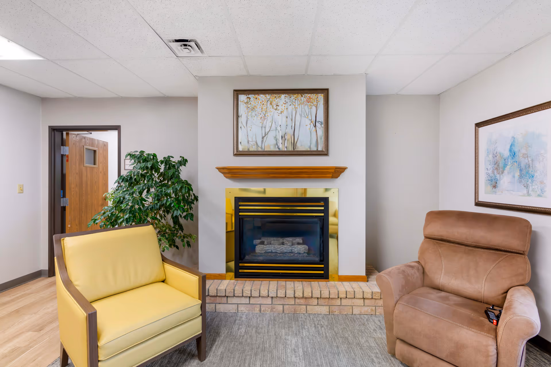 Cozy lounge area with a fireplace centered between a yellow armchair and a brown recliner, a potted plant and framed artwork.