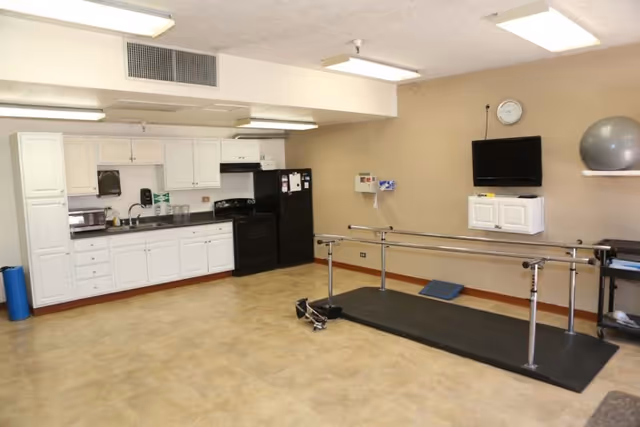 Spacious rehabilitation room with a kitchenette on the left, parallel walking bars on a mat in the center, a wall-mounted TV, and exercise equipment.