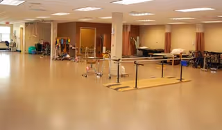 A spacious rehabilitation or physical therapy room with parallel bars for walking exercises, therapy beds, exercise equipment, and chairs along the walls. The room has a beige floor and ceiling lights.