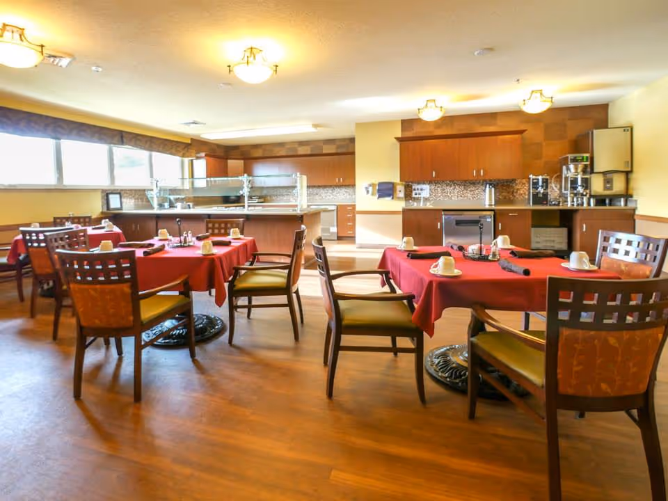 Dining room with several tables covered in red tablecloths, wooden chairs, place settings, and a serving counter/kitchen area in the background.