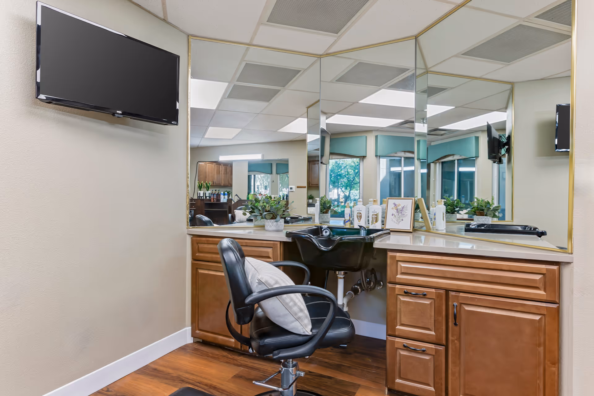 Interior view of a salon area with a black salon chair in front of a countertop with a built-in black sink. The countertop has various bottles and a framed picture. Large mirrors cover the walls behind the countertop, reflecting the room and windows with green valances. A flat-screen TV is mounted on the beige wall to the left.