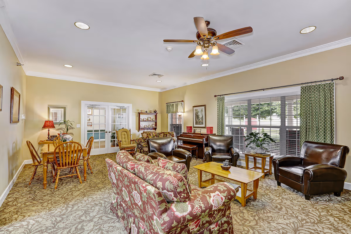 Bright communal living room with patterned sofas, leather armchairs, a coffee table, a dining set, and a piano by a large window.