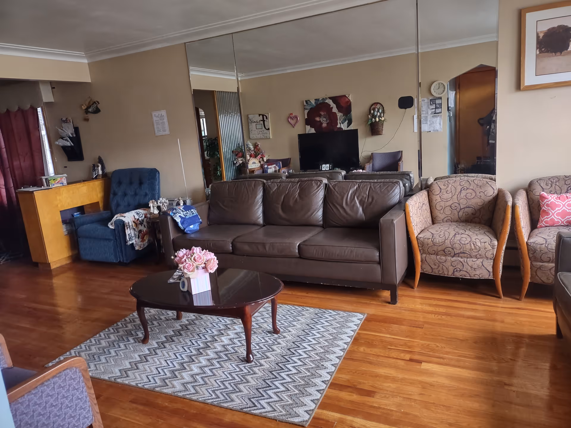 A cozy living room with a brown leather sofa, two patterned armchairs, a blue recliner, and a wooden coffee table with a flower arrangement on a patterned rug. The room has hardwood floors, beige walls, a large mirror on one wall, and a TV with floral decorations behind it.