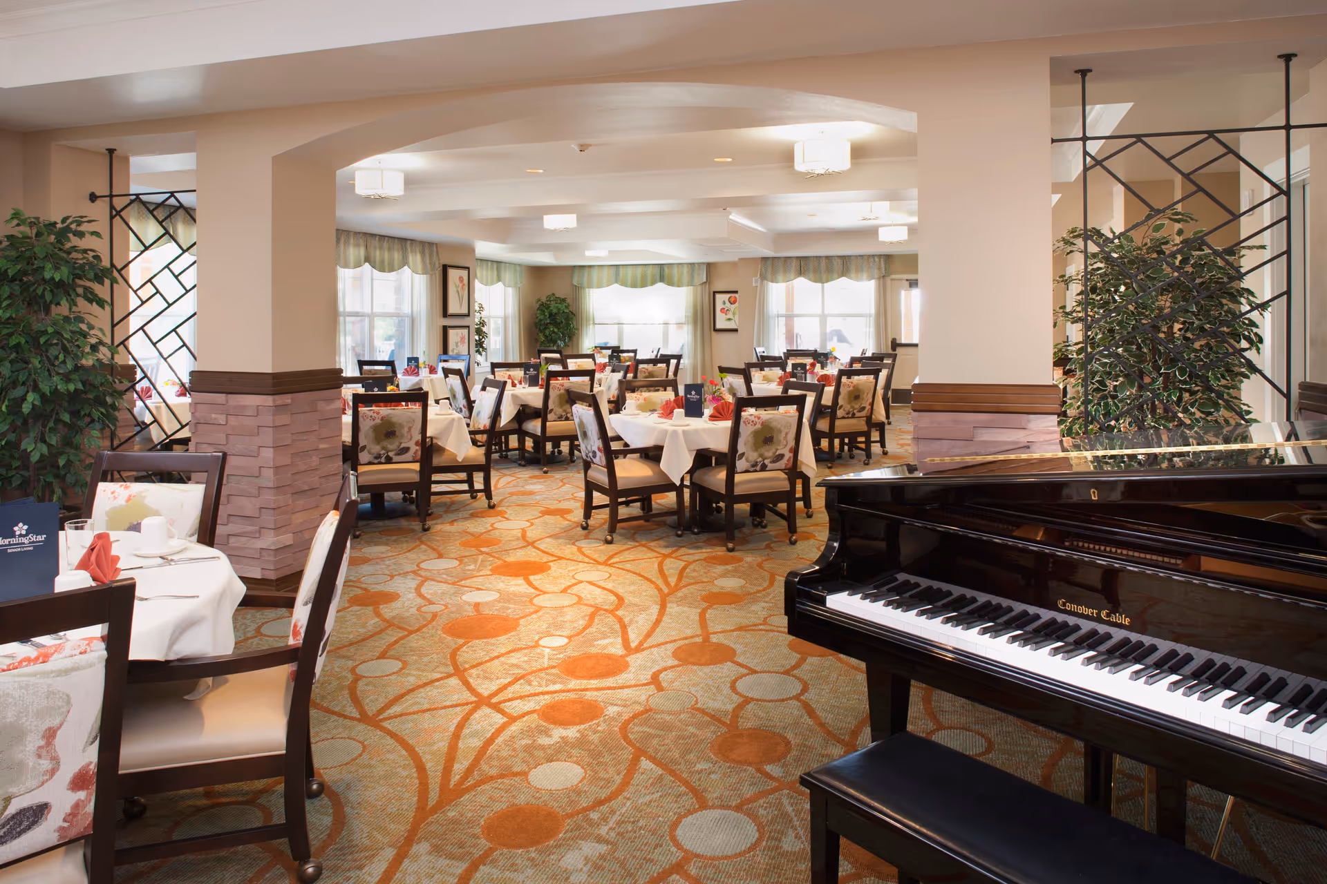 A bright and spacious dining room with multiple tables covered in white tablecloths and set with napkins and tableware. The room features floral upholstered chairs, large windows with green valances, potted plants, and a black grand piano in the foreground on the right side. The carpet has an orange and beige pattern, and the walls are painted in neutral tones with decorative columns.