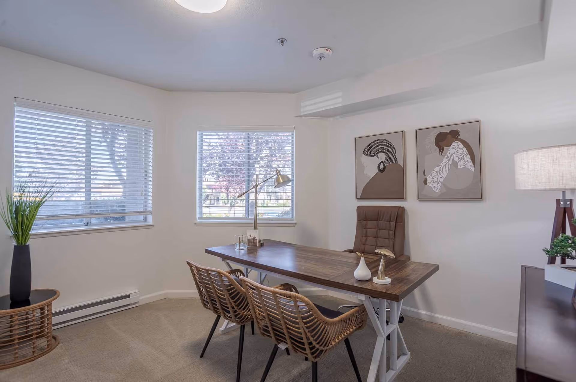 A bright office space with two large windows featuring white blinds. The room has a wooden desk with a brown cushioned chair behind it and two wicker chairs in front. On the desk are decorative items including a white vase and a gold lamp. The walls are white with two framed abstract art pieces. A side table with a tall black vase holding green plants is in the corner, and a floor lamp with a beige shade is next to a dark wooden cabinet.