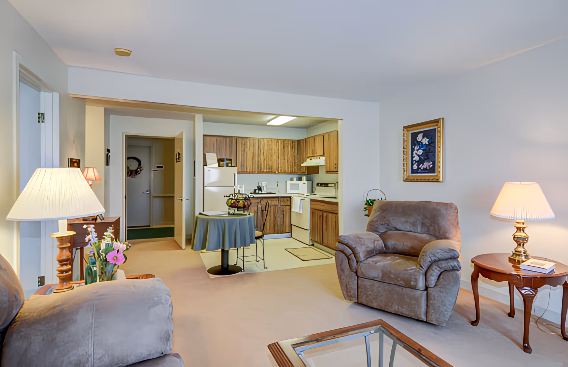 A cozy living room area with two brown upholstered armchairs, a wooden side table with a lamp and a book, and a glass coffee table. The room opens into a small kitchen with wooden cabinets, a white refrigerator, stove, and microwave. A small round table with a blue tablecloth and a chair is positioned between the living room and kitchen. The walls are light-colored, and there is a framed artwork hanging on the wall.