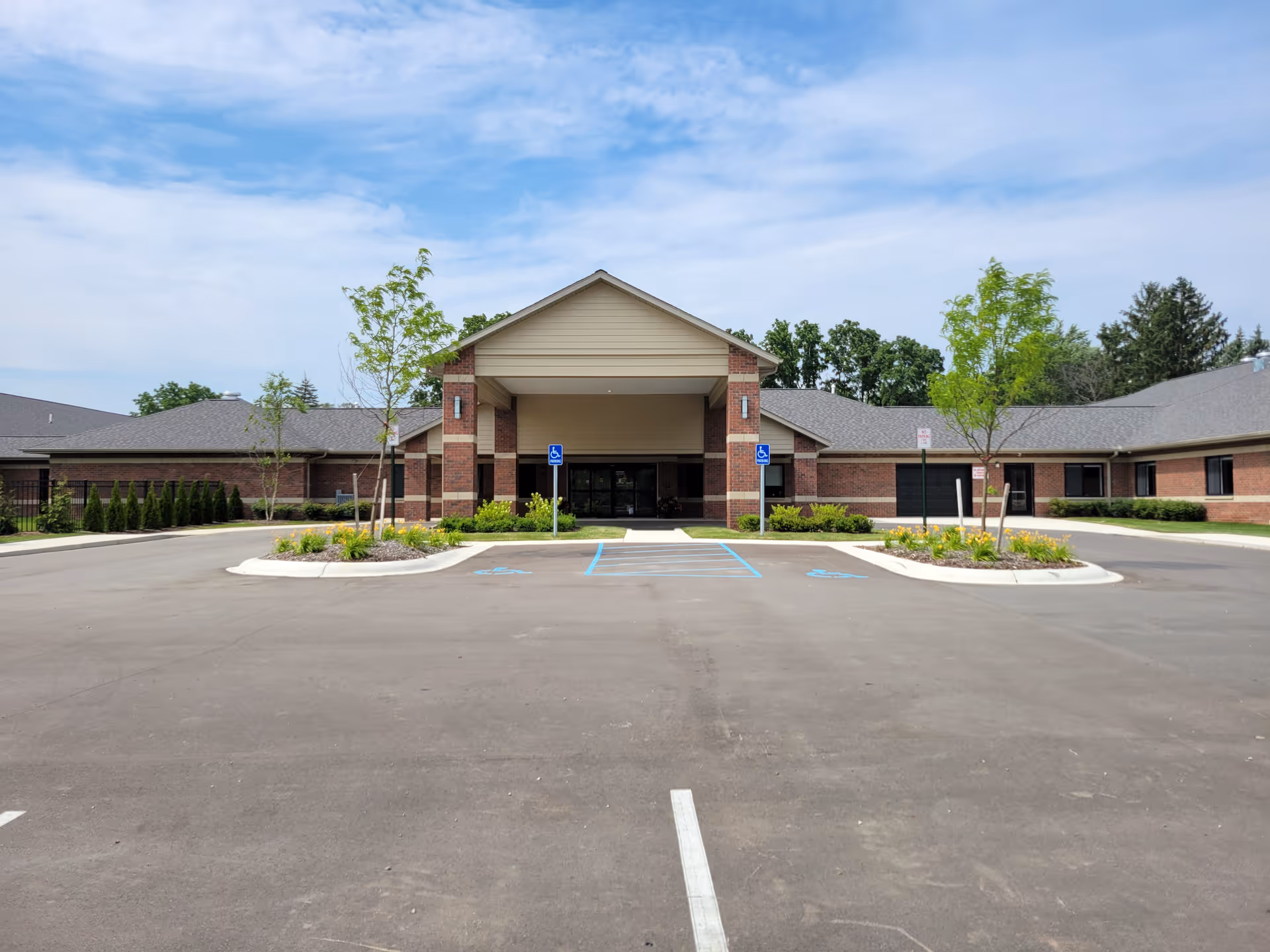Front entrance of a single-story brick assisted living building with a covered porte-cochere, landscaped islands, and marked handicapped parking spaces under a blue sky.