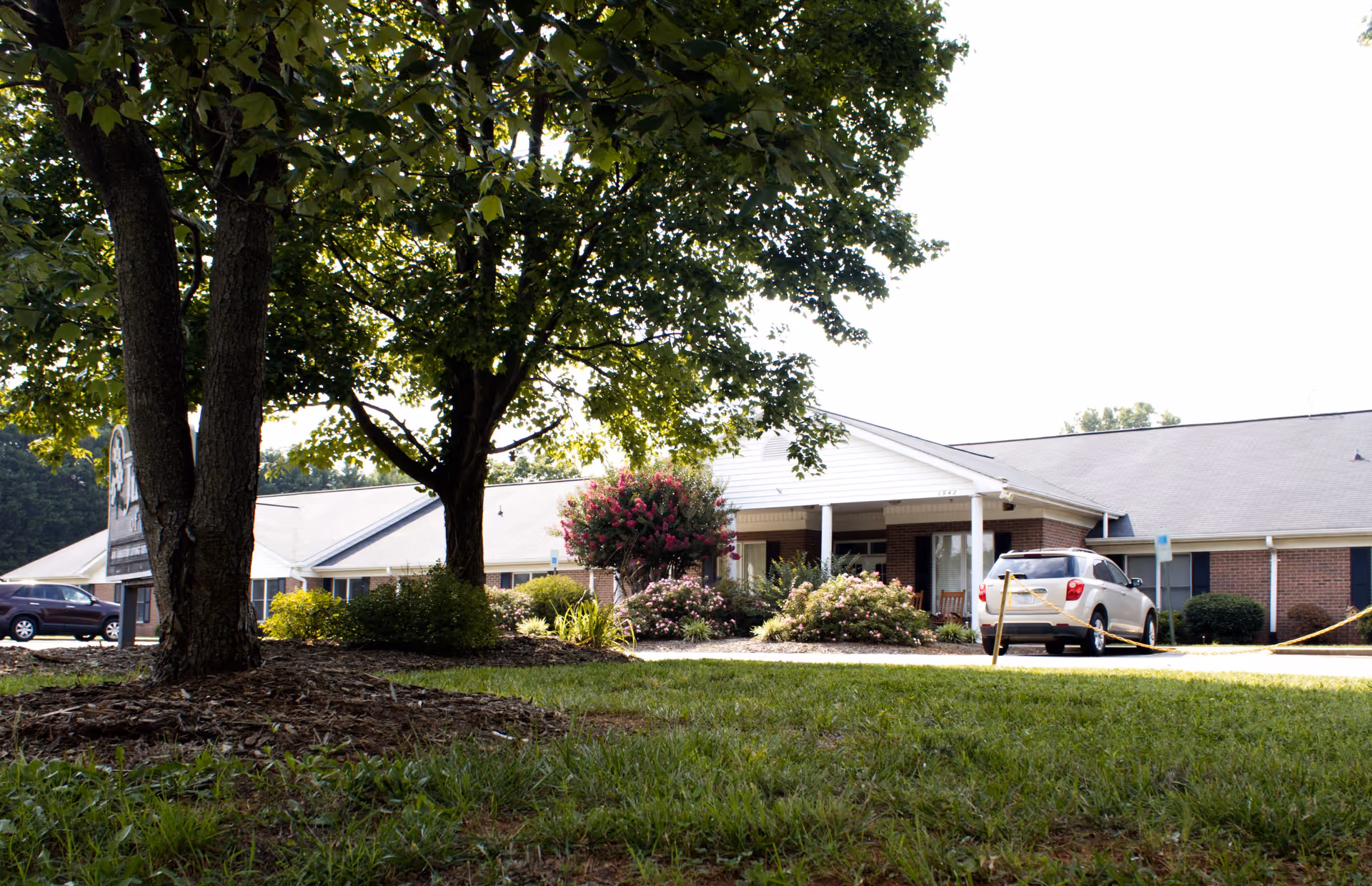 Exterior view of Jurney's of Statesville facility showing a single-story brick building with white trim, a covered entrance, landscaped bushes and flowering plants, a tree in the foreground, and parked cars in the driveway.