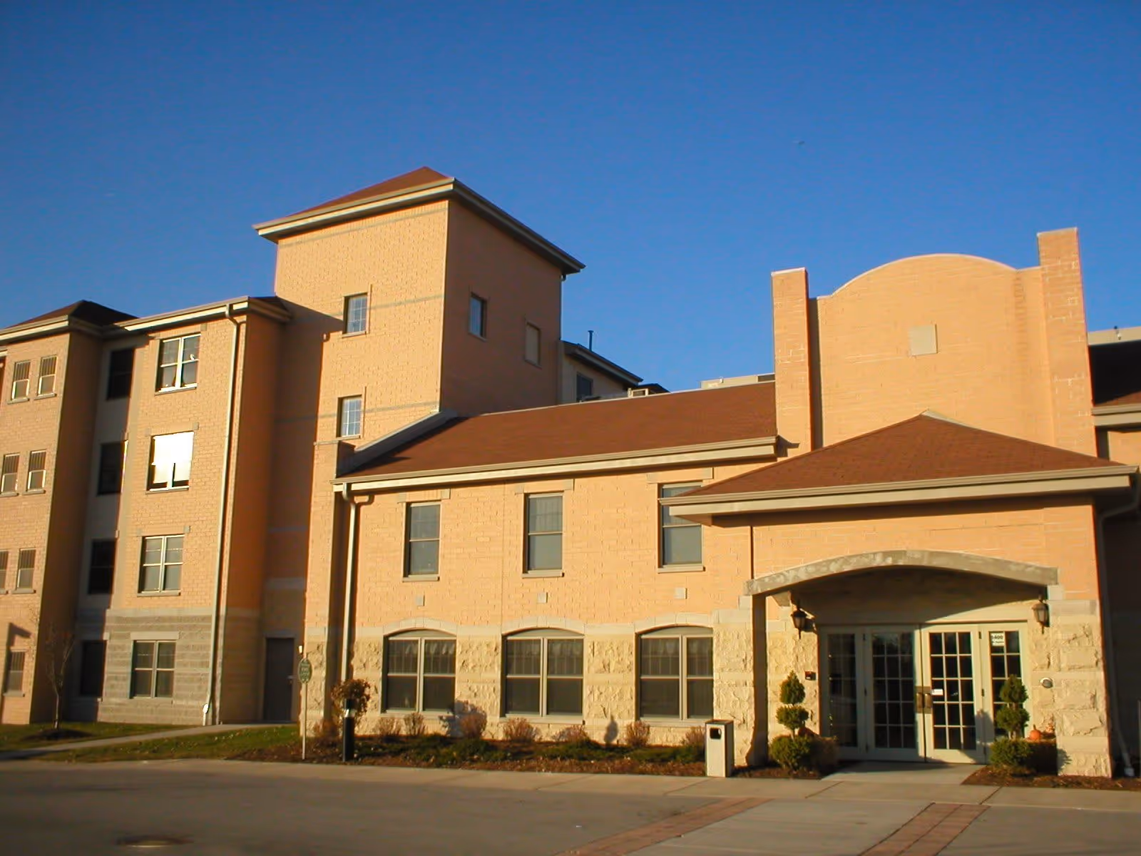 Exterior view of a multi-story senior living facility building with beige brick walls, multiple windows, and a main entrance with glass doors under a small roof. The sky is clear and blue.