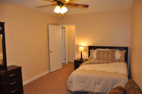A bedroom with beige walls and carpeted floor featuring a bed with a dark wooden headboard, beige bedding, and patterned pillows. There is a nightstand with a lamp beside the bed, a dresser with a mirror on the left side, and a ceiling fan with lights overhead. The door to the room is open, showing a hallway with additional doors.