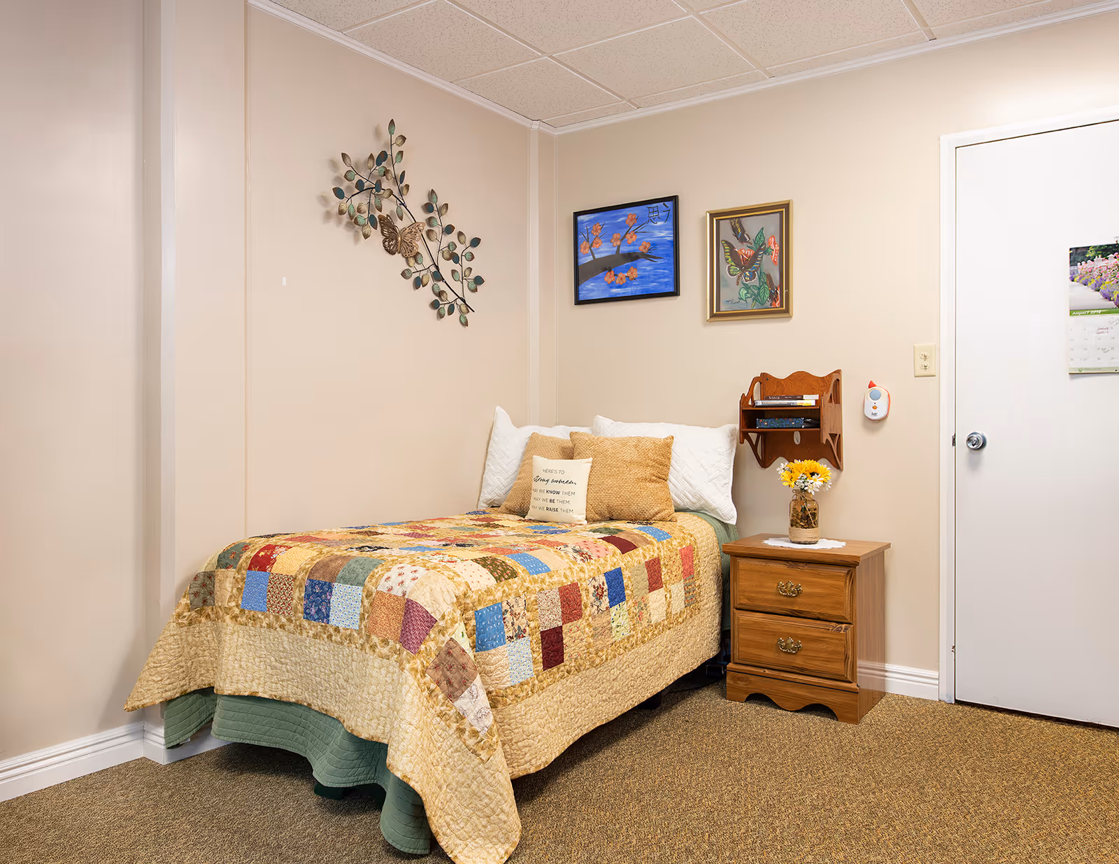 Neat single bedroom with a colorful patchwork quilted bed, wooden nightstand with flowers, and wall art above the bed.