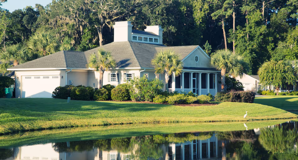 A single-story building with a gray roof and white exterior surrounded by palm trees and bushes, reflected in a calm pond in the foreground, with a white bird standing near the water's edge and dense trees in the background.