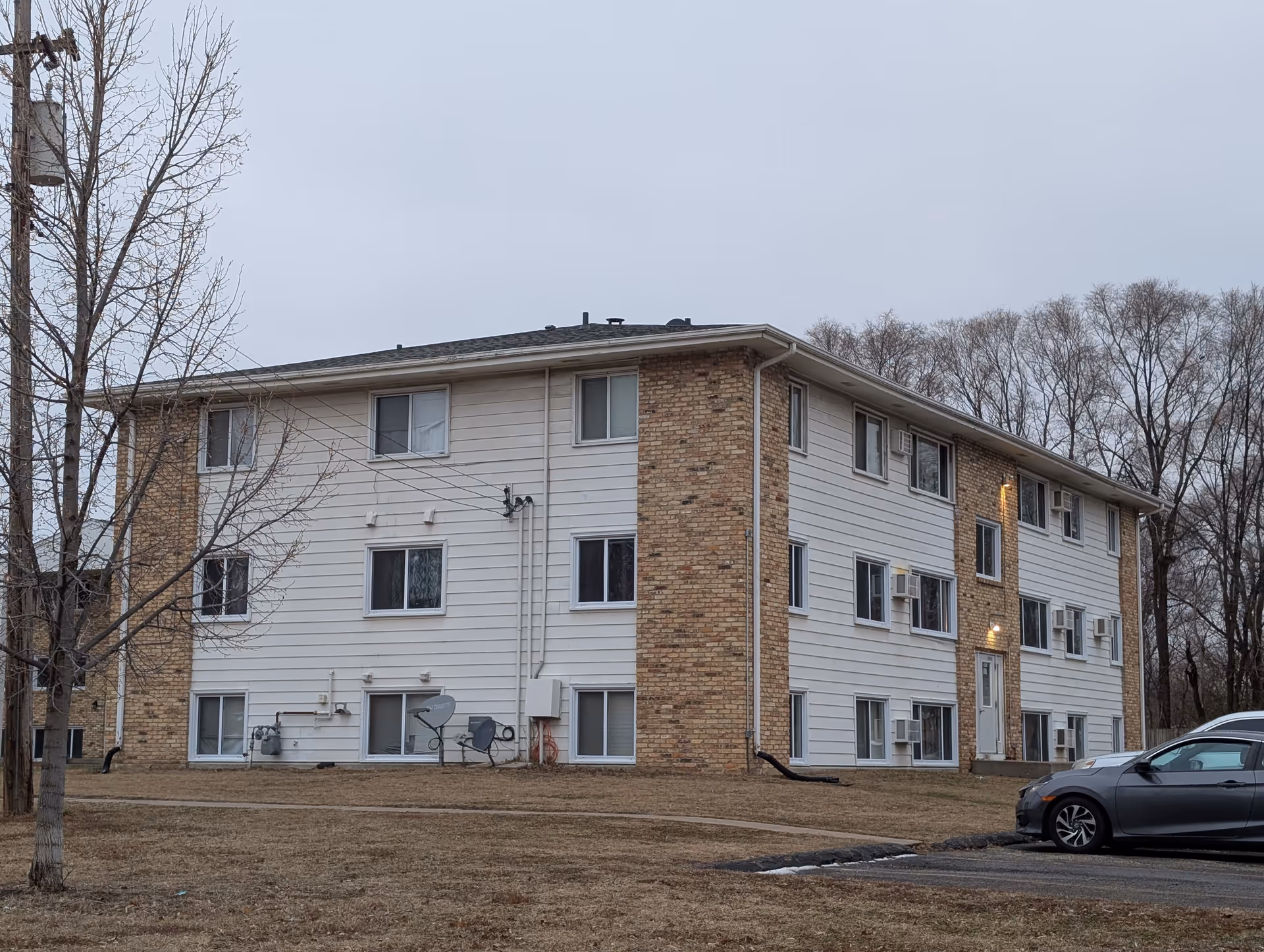 Three-story brick-and-white-siding apartment building with multiple windows, satellite dishes, and a parked car on a grassy lot.