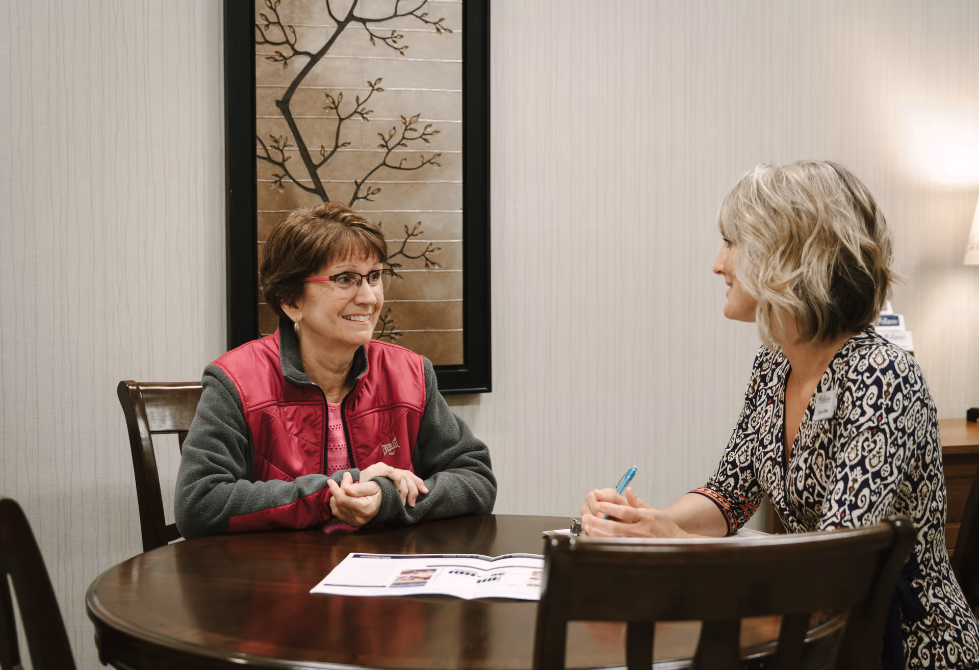 Two women sitting at a round wooden table in a room with beige wallpaper and a decorative wall art featuring a tree branch. One woman is wearing glasses and a red vest, smiling, while the other woman, with blonde hair and a patterned blouse, is holding a pen and appears to be engaged in conversation.