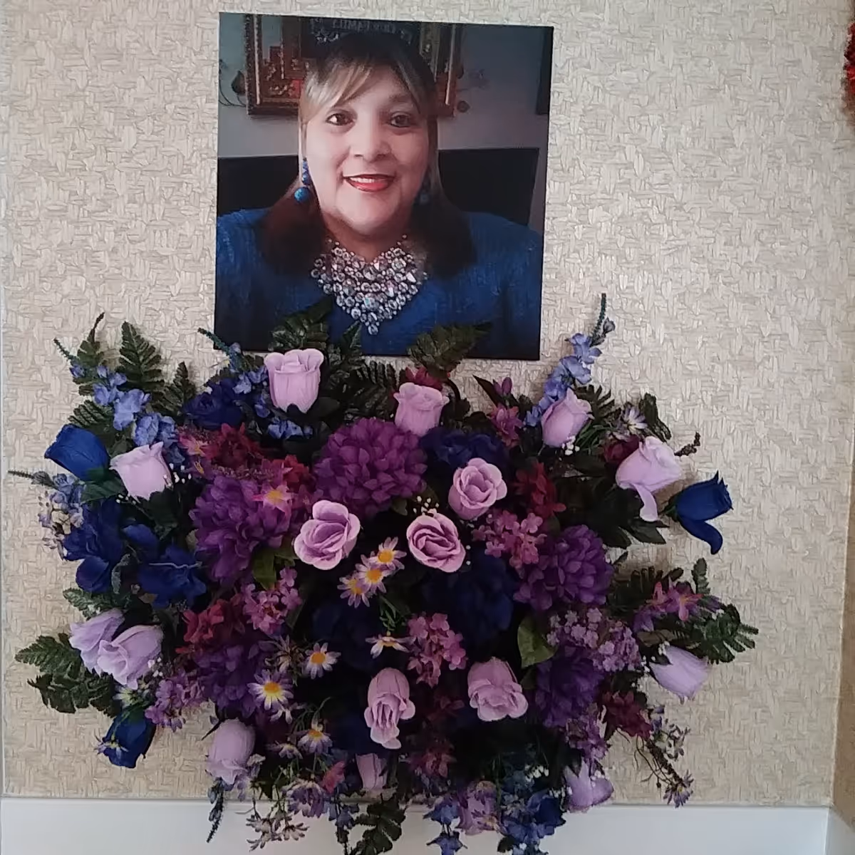 A framed portrait of a smiling woman wearing a blue top and a large decorative necklace, hanging on a textured beige wall above a large bouquet of purple, lavender, and blue flowers with green leaves.