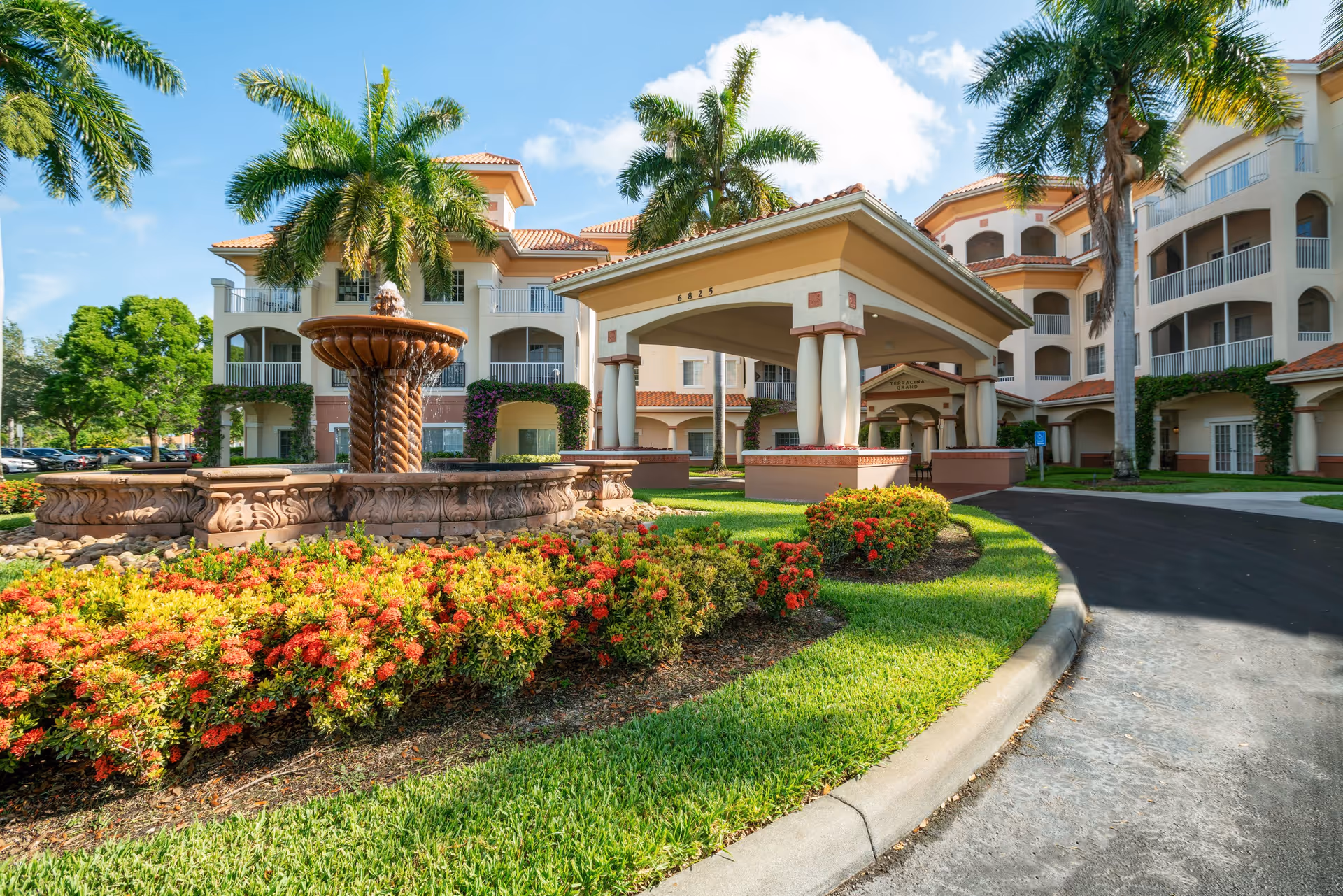 Exterior view of Terracina Senior Living facility featuring a large covered entrance with columns, a decorative fountain surrounded by colorful flowers and greenery, palm trees, and a multi-story building with balconies under a blue sky with some clouds.