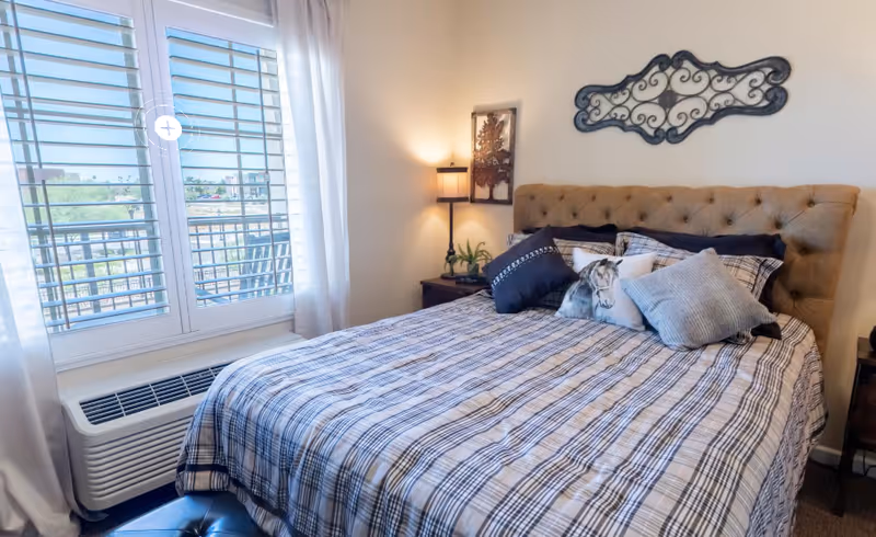 Sunlit bedroom featuring a tufted headboard, plaid bedding with decorative pillows, nightstands and a shuttered window.