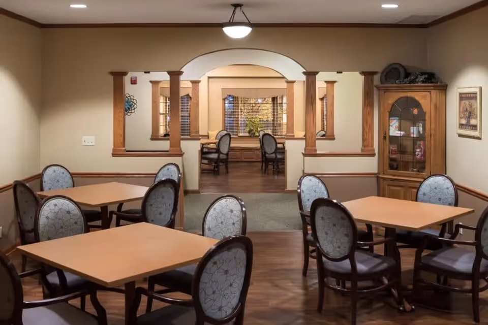 A cozy dining area in a senior living facility with several square wooden tables and cushioned chairs with floral patterned upholstery. The room features warm beige walls with wooden trim, a wooden floor, and a wooden cabinet with glass doors displaying decorative items. An arched opening with wooden columns leads to another room with additional seating and windows with blinds.