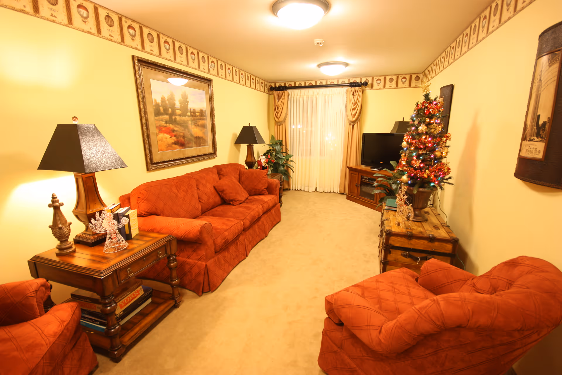 Cozy living room with red upholstered sofas and chairs, side tables and lamps, a small decorated Christmas tree, TV, and framed artwork.
