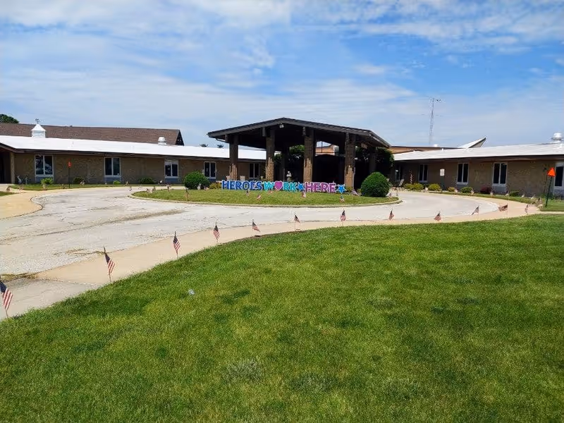 Front entrance of a single-story senior living facility with a covered porte-cochère, circular driveway, lawn and a sign reading 'HEROES WORK HERE'.