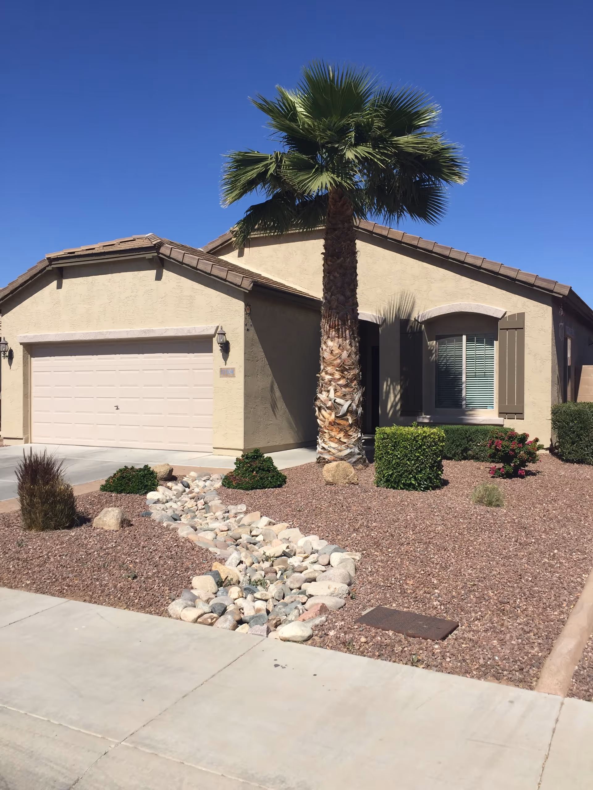 Exterior view of a single-story beige stucco house with a tiled roof, a two-car garage, a palm tree, and desert landscaping with rocks and shrubs under a clear blue sky.