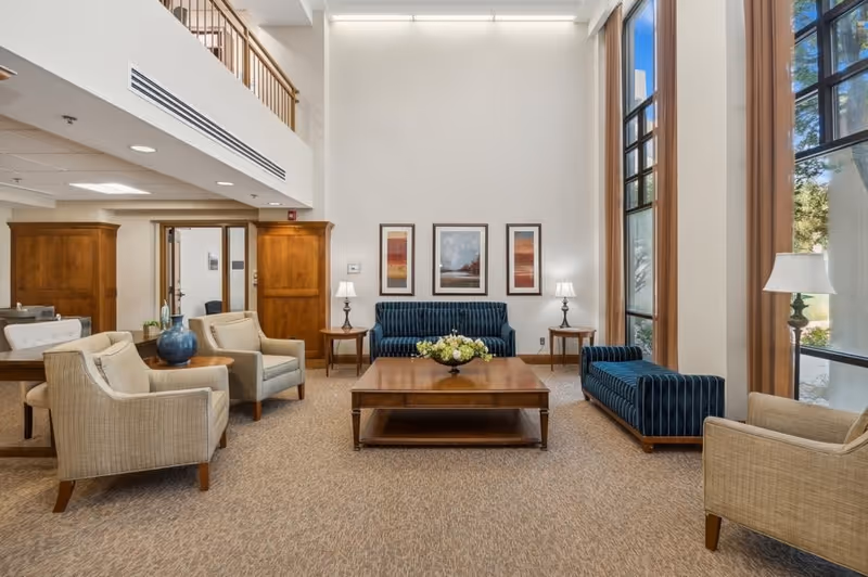 Bright, spacious senior living common room with sofas, armchairs, a central coffee table, tall windows, and framed artwork on the wall.