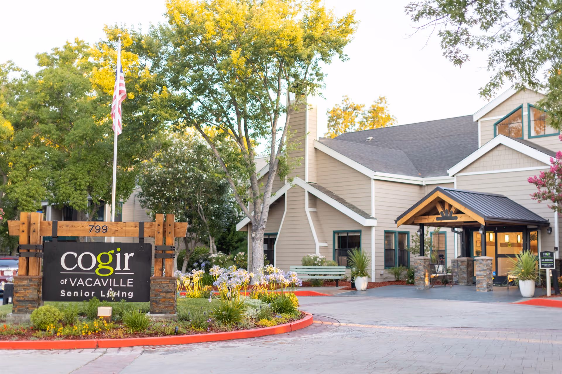 Exterior view of Cogir of Vacaville Senior Living facility showing the building entrance with a covered porch, surrounded by trees and landscaping. A sign with the facility name is prominently displayed near the driveway, along with an American flag on a flagpole.