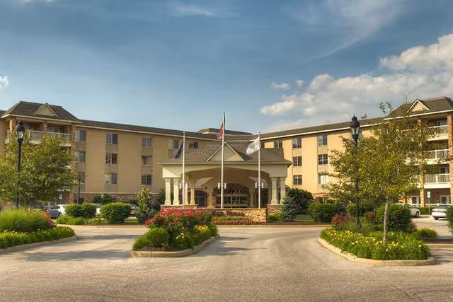 Front exterior view of Westside Garden Plaza, a multi-story senior living facility with a covered entrance, three flagpoles, landscaped greenery, and a circular driveway under a partly cloudy sky.