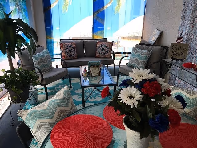 A cozy indoor seating area with a glass-top table and several cushioned chairs and a loveseat. The cushions have blue and white chevron and patterned designs. A vase with red, white, and blue flowers sits on the table, along with red placemats. The background features a large window with a tropical beach scene curtain, and there are some decorative items including a 'Give Thanks' sign and small American flags.
