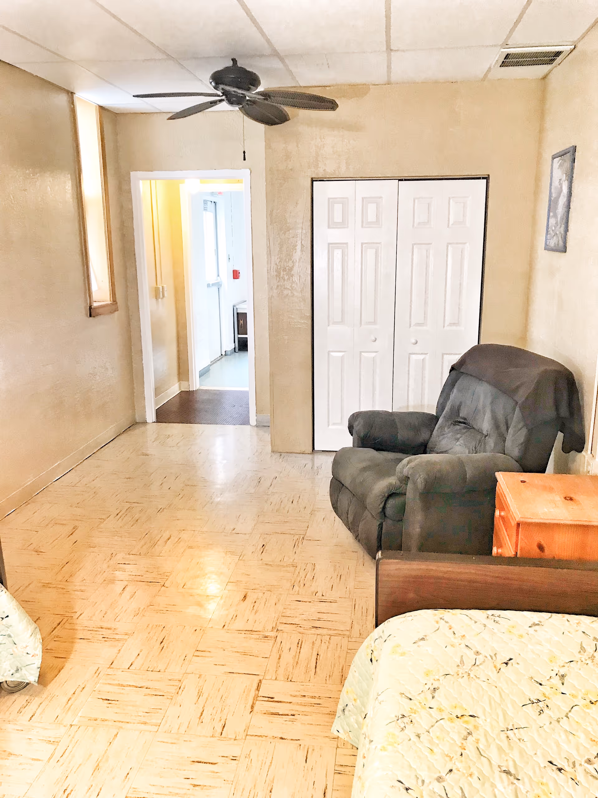 Small assisted-living bedroom with a recliner, bed corner, nightstand, closet doors and a ceiling fan.