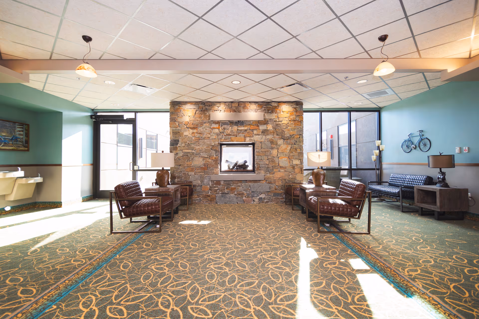 A bright and spacious lounge area with a stone fireplace in the center. The room features patterned carpet, two pairs of brown leather chairs with side tables and lamps, a black cushioned bench, and decorative elements including a wall-mounted bicycle and a painting. Large windows allow natural light to fill the space.