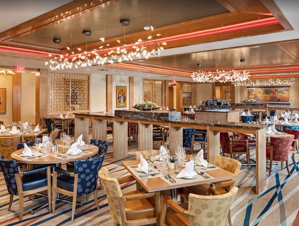 A spacious, elegantly decorated dining room with multiple round and square tables set with white napkins, glassware, and silverware. The room features patterned chairs in blue, red, and yellow, modern chandeliers with leaf-like light fixtures, and decorative wall art. The ceiling has recessed lighting with red accents, and the floor is covered with a striped carpet.