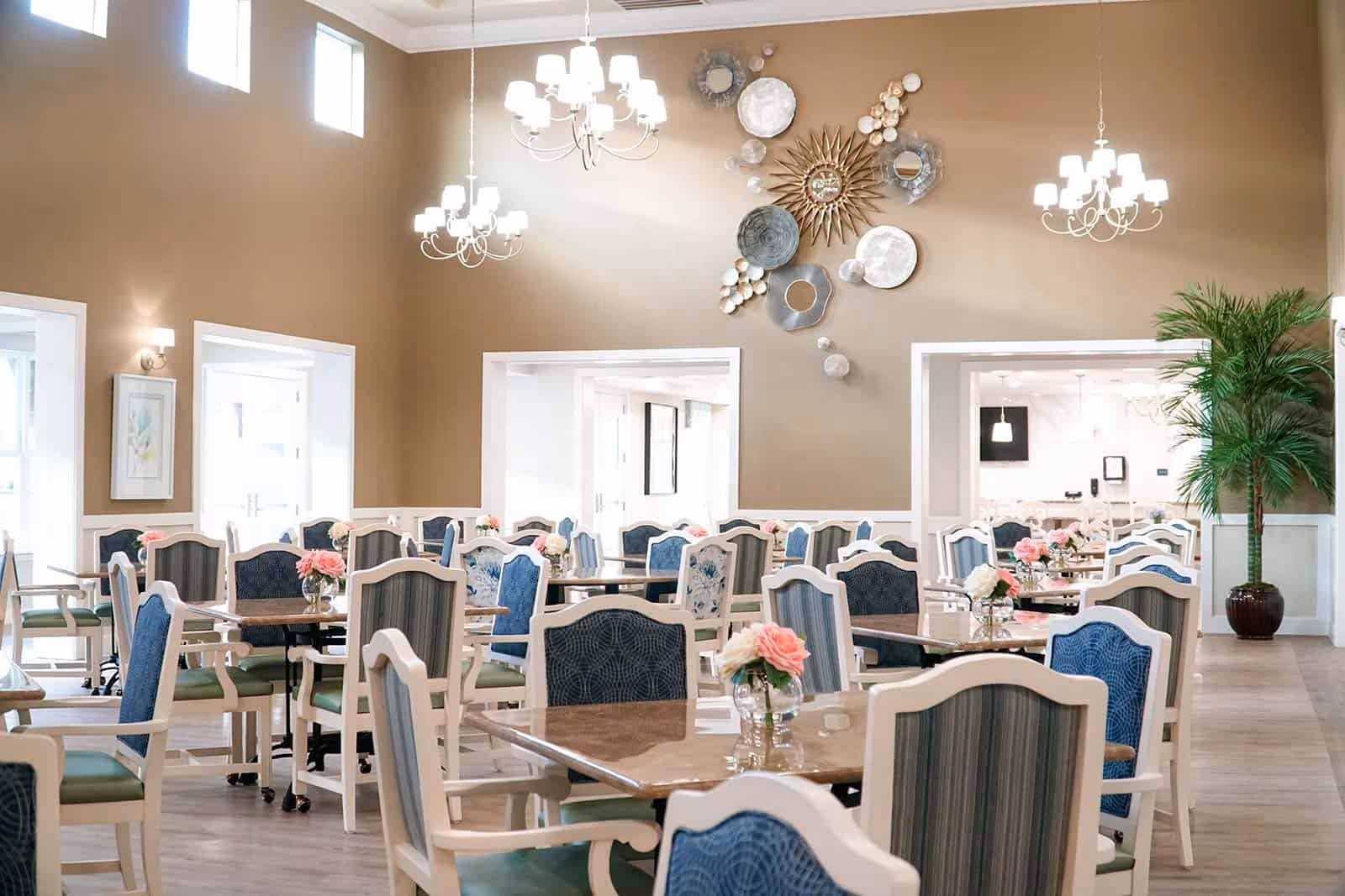 A bright and spacious dining room with multiple tables and chairs arranged neatly. Each table has a small vase with pink and white flowers. The walls are painted beige with white trim, and there are three chandeliers hanging from the high ceiling. Decorative wall art featuring circular metallic designs is displayed on one wall, and a large potted plant is placed in the corner.