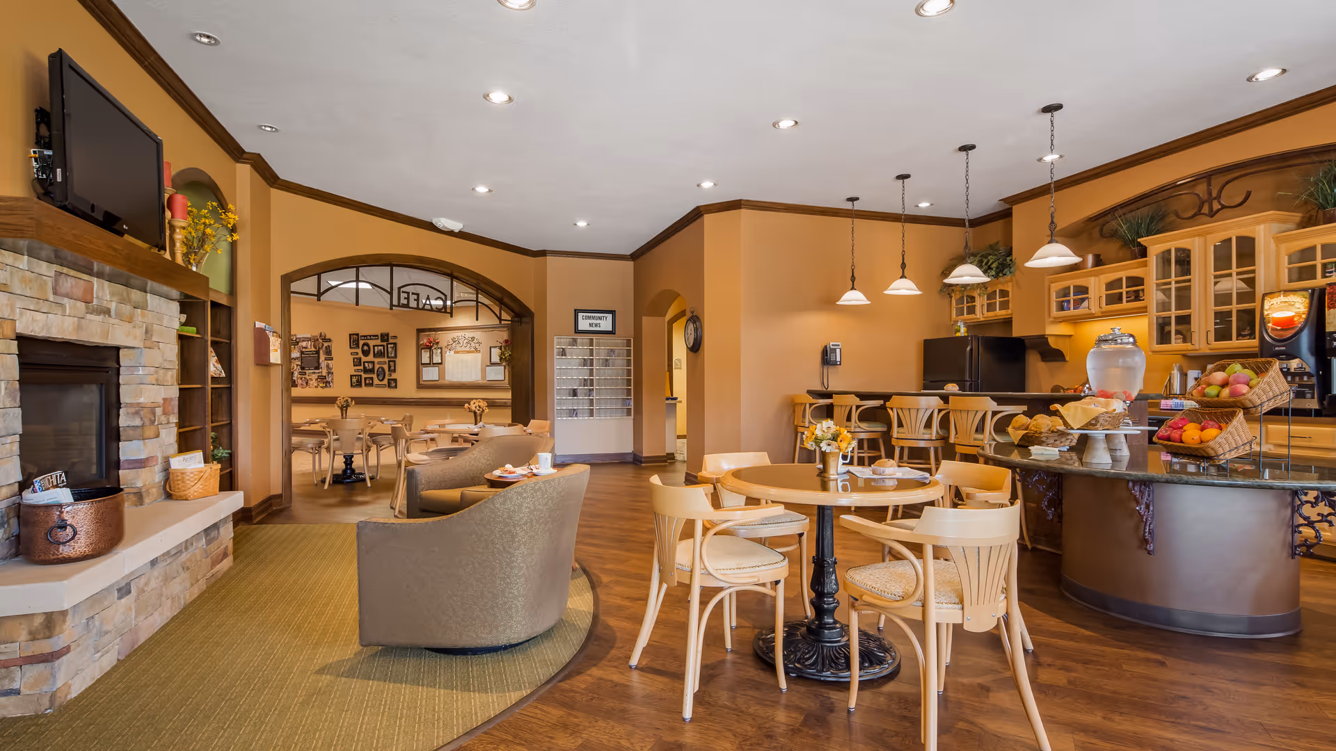 A warm and inviting common area in a senior living facility featuring a stone fireplace with a mounted TV above it, comfortable armchairs, round wooden tables with chairs, and a kitchen area with hanging pendant lights, a refrigerator, and a counter displaying baskets of fruit and baked goods. In the background, there is an archway leading to a dining area with more tables and chairs, and a community news board on the wall.