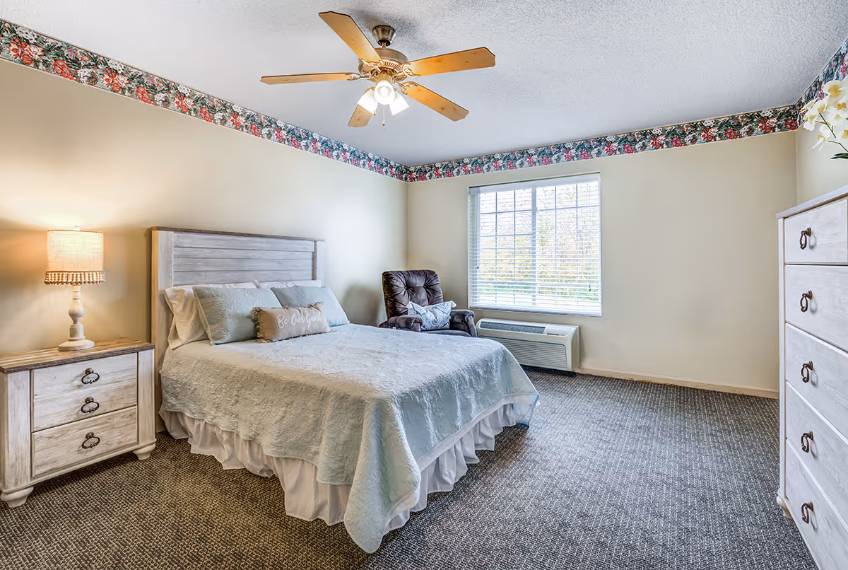 A bedroom with a large bed covered in a light blue quilt and multiple pillows, including one with the words 'Be Our Guest'. There is a wooden nightstand with a lamp on the left side of the bed, a cushioned armchair near a window with blinds, and a wooden dresser on the right side. The room has beige walls with floral wallpaper border near the ceiling and a ceiling fan with lights.